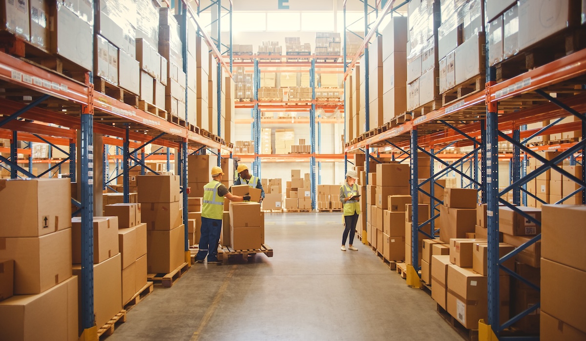 段ボール箱に入った商品が棚いっぱいに並ぶ小売倉庫で、作業員が箱をスキャンして仕分けし、パレットトラックやフォークリフトで在庫を移動している。製品流通配送センター。