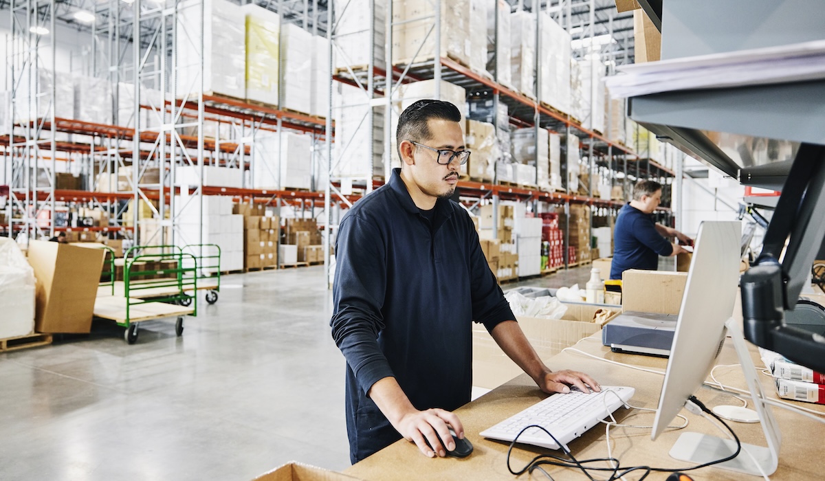 Medium wide shot of male warehouse worker checking orders at computer workstation in warehouse
