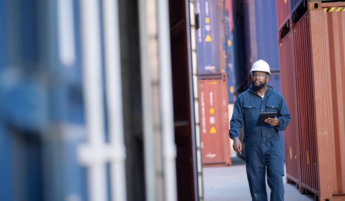 Technology is Helping Ports Optimize Their operations in Global Containerization Services. African American male transport engineer walking with holding a tablet computer to examine cargo in a commercial dock.